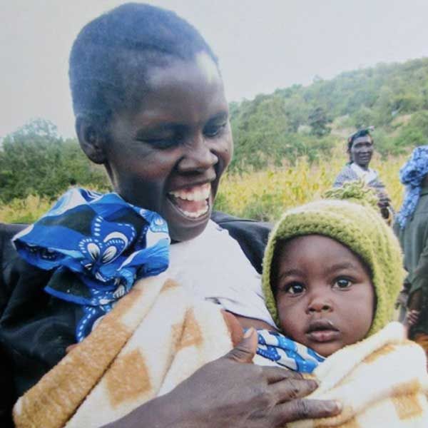 Woman laughing as she holds a baby wrapped in a blanket, with tall crops in the field behind them.