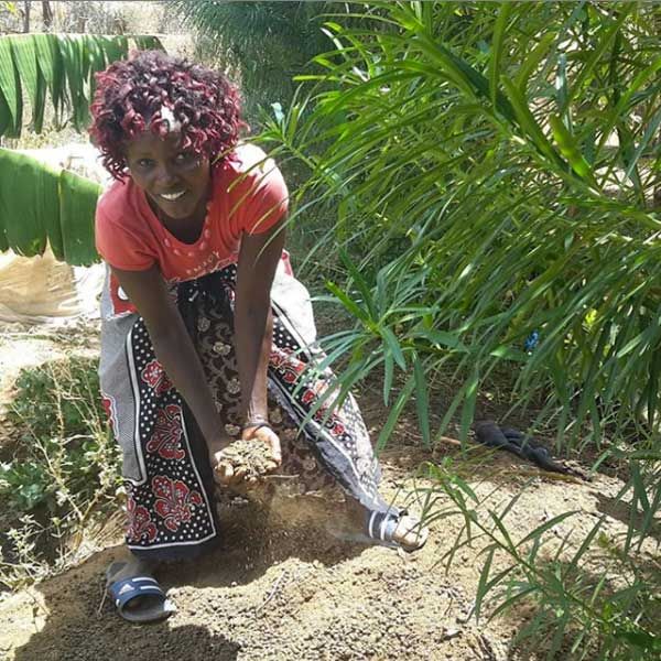Woman kneeling in a garden, lifting a handful of soil towards the camera.